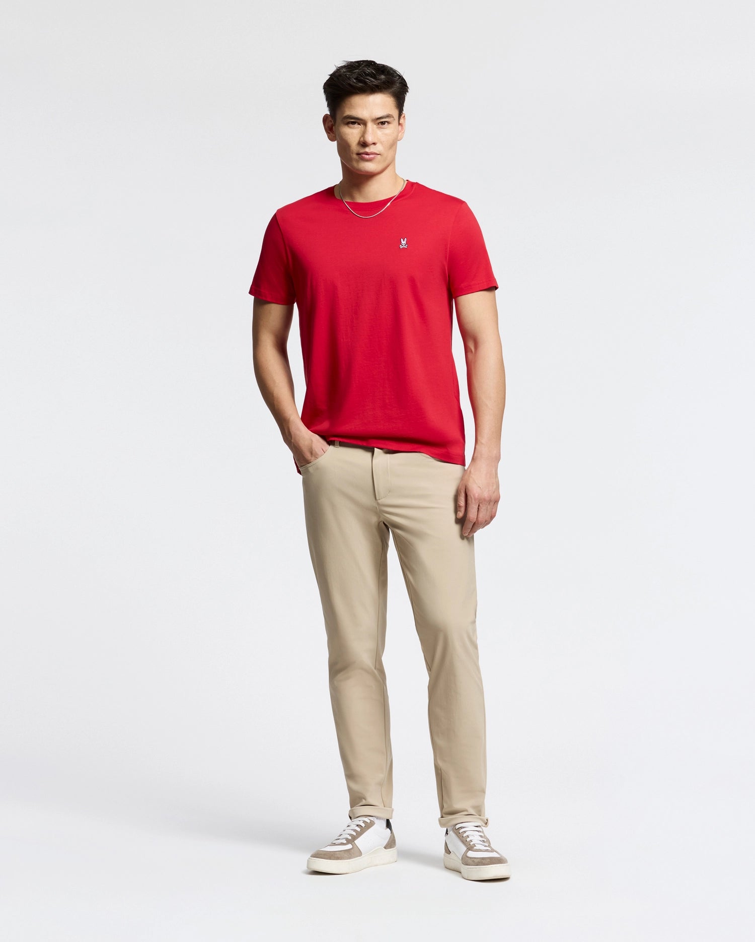 A person stands against a plain background wearing a Psycho Bunny MENS CLASSIC CREW NECK TEE in red, paired with beige pants and white sneakers. They have short dark hair, pose with hands in pockets, and look towards the camera.