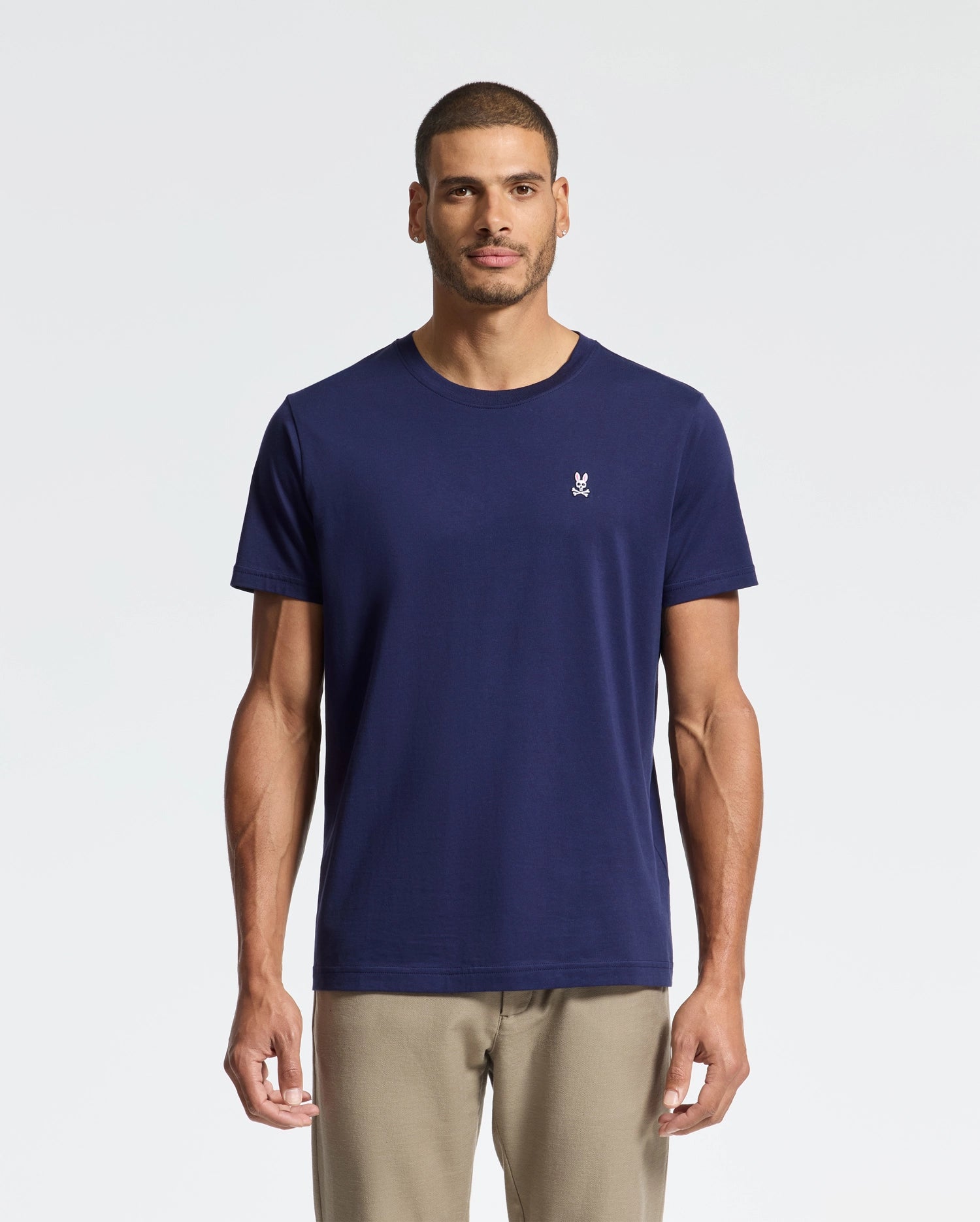 Against a white background, a man wears Psycho Bunny's navy blue "MENS CLASSIC CREW NECK TEE - B6U014CRPC" in soft Pima cotton and light beige pants. Facing the camera neutrally, his regular fit tee sports a small white logo on the left chest.
