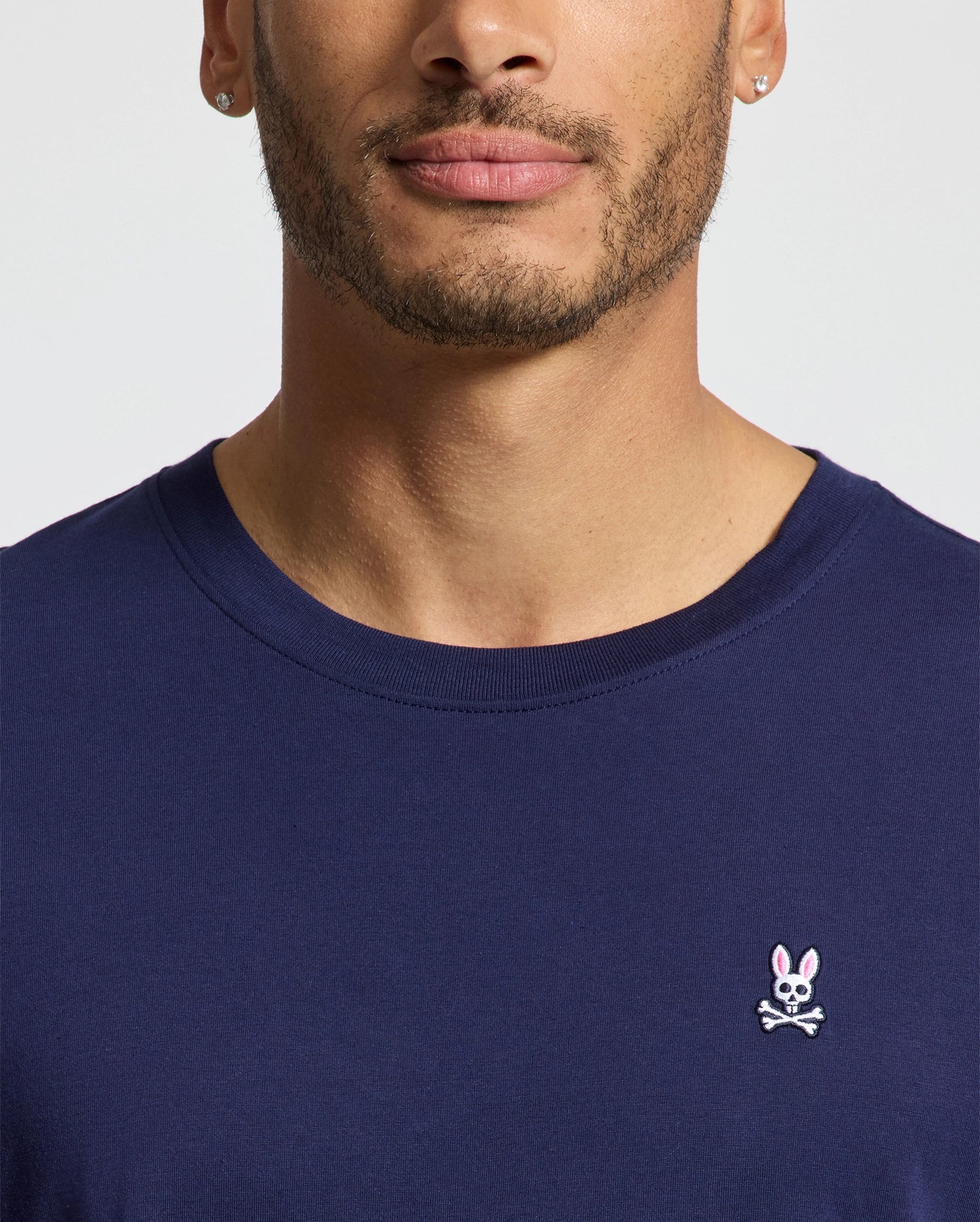 A close-up shows a person in a Psycho Bunny MENS CLASSIC CREW NECK TEE - B6U014CRPC in navy, featuring an embroidered pink rabbit skull and crossbones on the chest, made from soft pima cotton. The regular fit tee complements their short facial hair, visible from shoulders up.