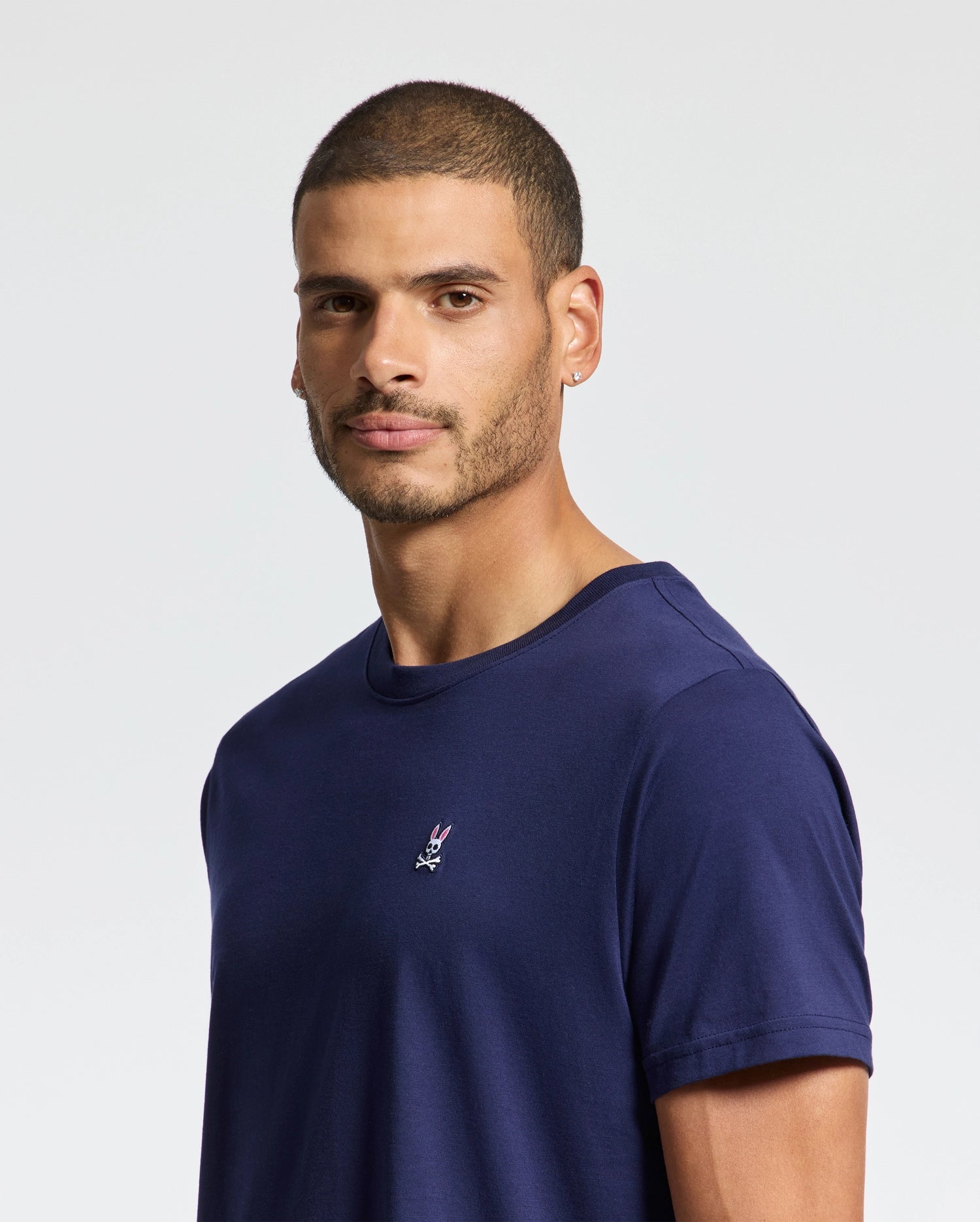 A man with short hair and a beard wears a Psycho Bunny navy blue MENS CLASSIC CREW NECK TEE - B6U014CRPC, made of soft pima cotton. The regular fit shirt features a small embroidered emblem on the chest, enhancing his look as he gazes at the camera against a plain white background.