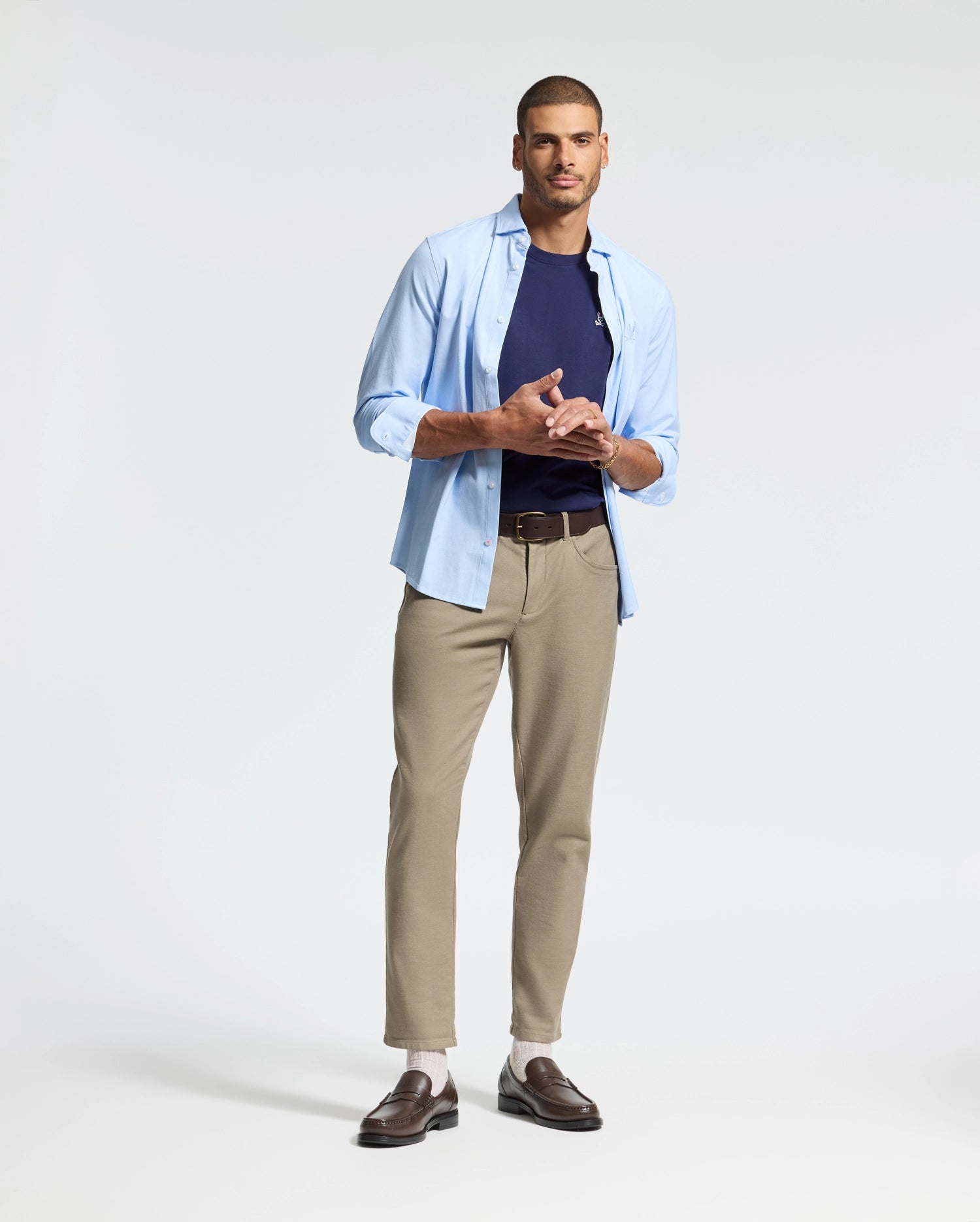 A man in a light blue, regular-fit shirt with rolled-up sleeves, wearing a Psycho Bunny MENS CLASSIC CREW NECK TEE - B6U014CRPC underneath, beige pants, and brown loafers stands against a plain white background. He has short hair and rests his arms on his chest.