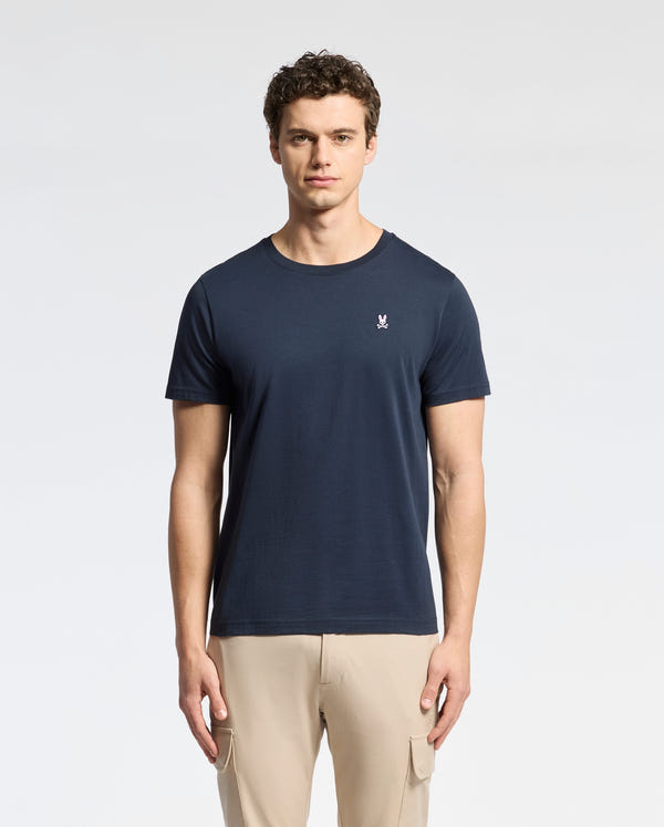 A man with short curly hair wears the Psycho Bunny MENS CLASSIC CREW NECK TEE - B6U014CRPC in navy blue and beige pants, standing against a plain light background and looking straight at the camera with a neutral expression.