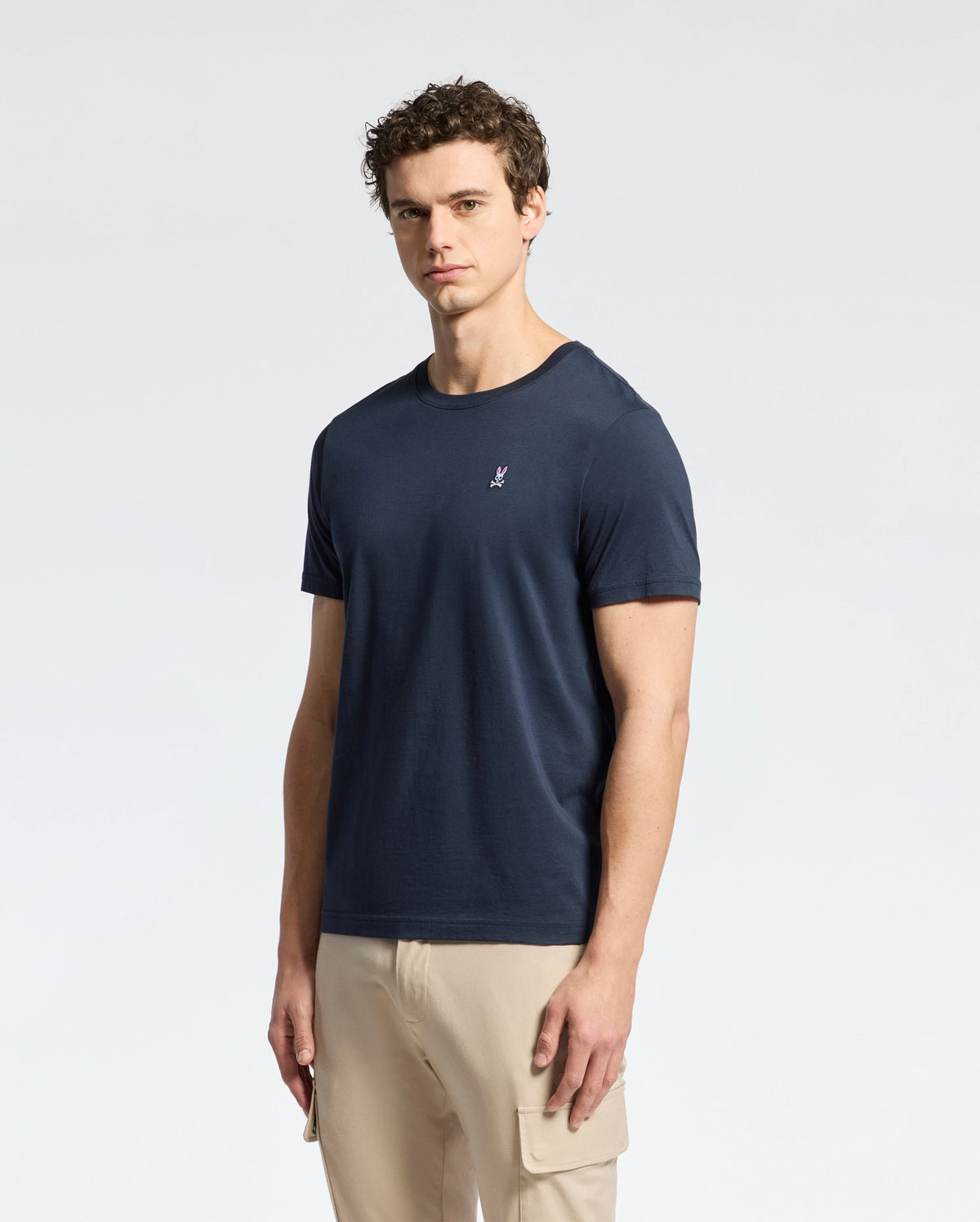 A man with short curly hair wears a Psycho Bunny MENS CLASSIC CREW NECK TEE - B6U014CRPC in navy blue, made from 100% Pima cotton, featuring a small chest logo, paired with beige cargo pants and standing against a light background.