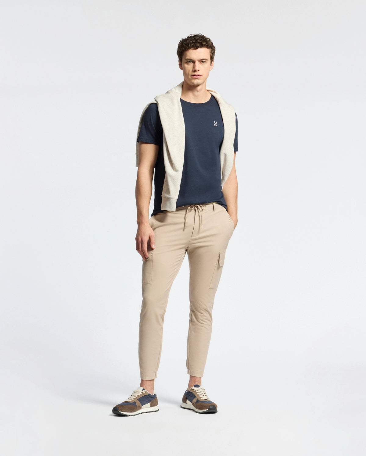 A man faces forward, wearing a Psycho Bunny MENS CLASSIC CREW NECK TEE - B6U014CRPC in navy (100% Pima cotton), paired with light beige joggers, multicolored sneakers, and a light gray sweater over his shoulders against a plain background.
