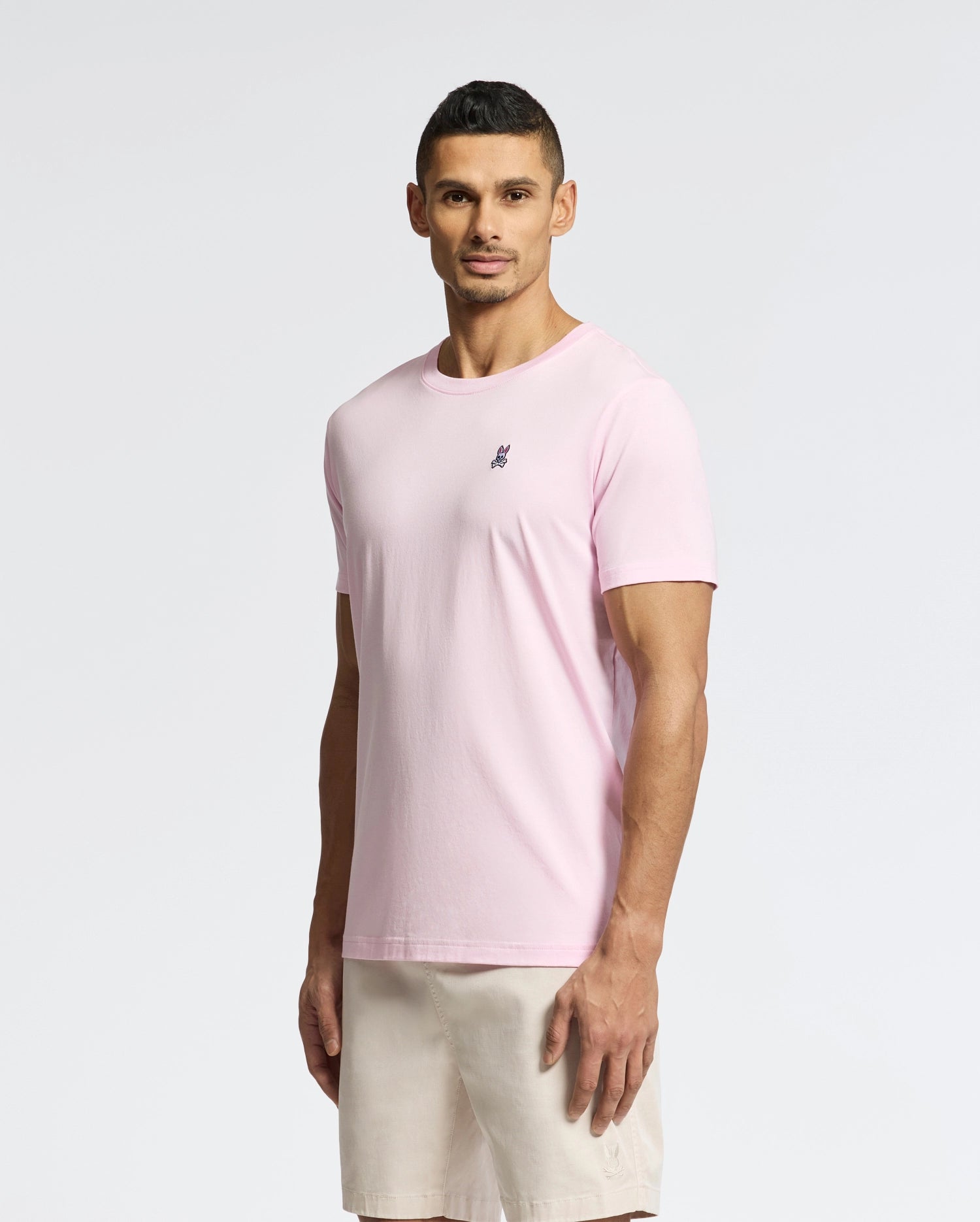 Against a plain backdrop, a person wears the Psycho Bunny Men's Classic Crew Neck Tee (B6U014CRPC) in light pink, made from soft pima cotton featuring a small chest emblem, paired with beige shorts. Its regular fit complements their slight smile as they gaze ahead.