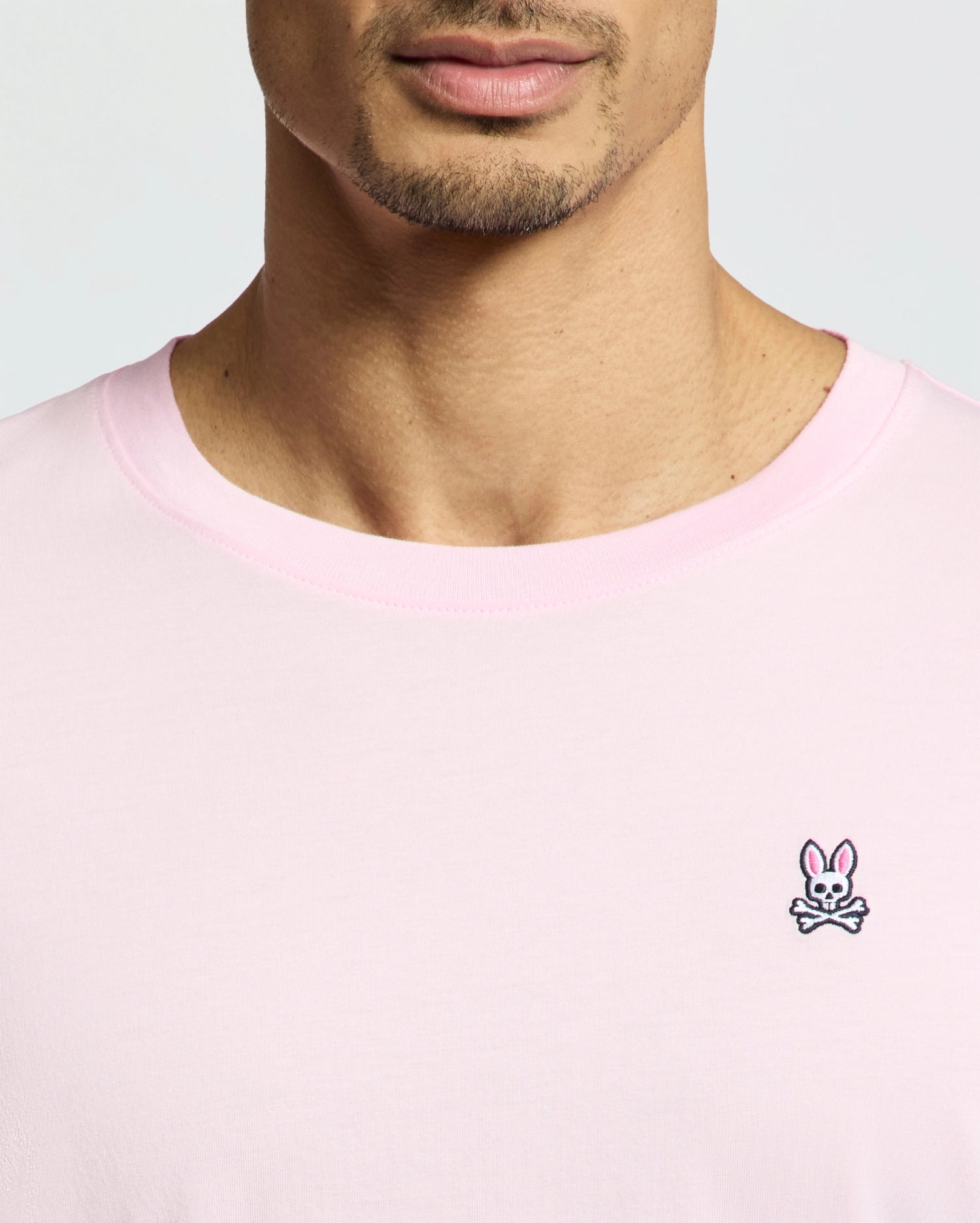 A close-up captures a person wearing the Psycho Bunny MENS CLASSIC CREW NECK TEE - B6U014CRPC in light pink. Made from soft pima cotton, it sports a small embroidered bunny with crossbones logo on the left. The regular fit tee shows their face partially from the nose down against a plain background.