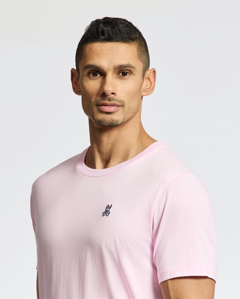 A man with short black hair and a trimmed beard wears the Psycho Bunny MENS CLASSIC CREW NECK TEE - B6U014CRPC in light pink, made from soft pima cotton with a small embroidered logo on the chest. He stands against a plain, light-toned background.