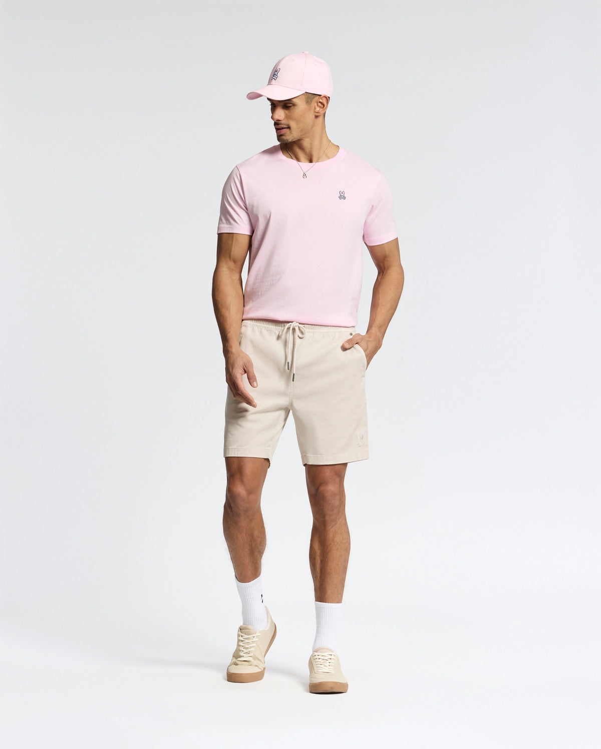 A man wears a light pink Psycho Bunny pima cotton cap and matching MENS CLASSIC CREW NECK TEE - B6U014CRPC, paired with beige drawstring shorts, white socks, and beige sneakers. Against a plain background, he looks down with his hands at his sides.