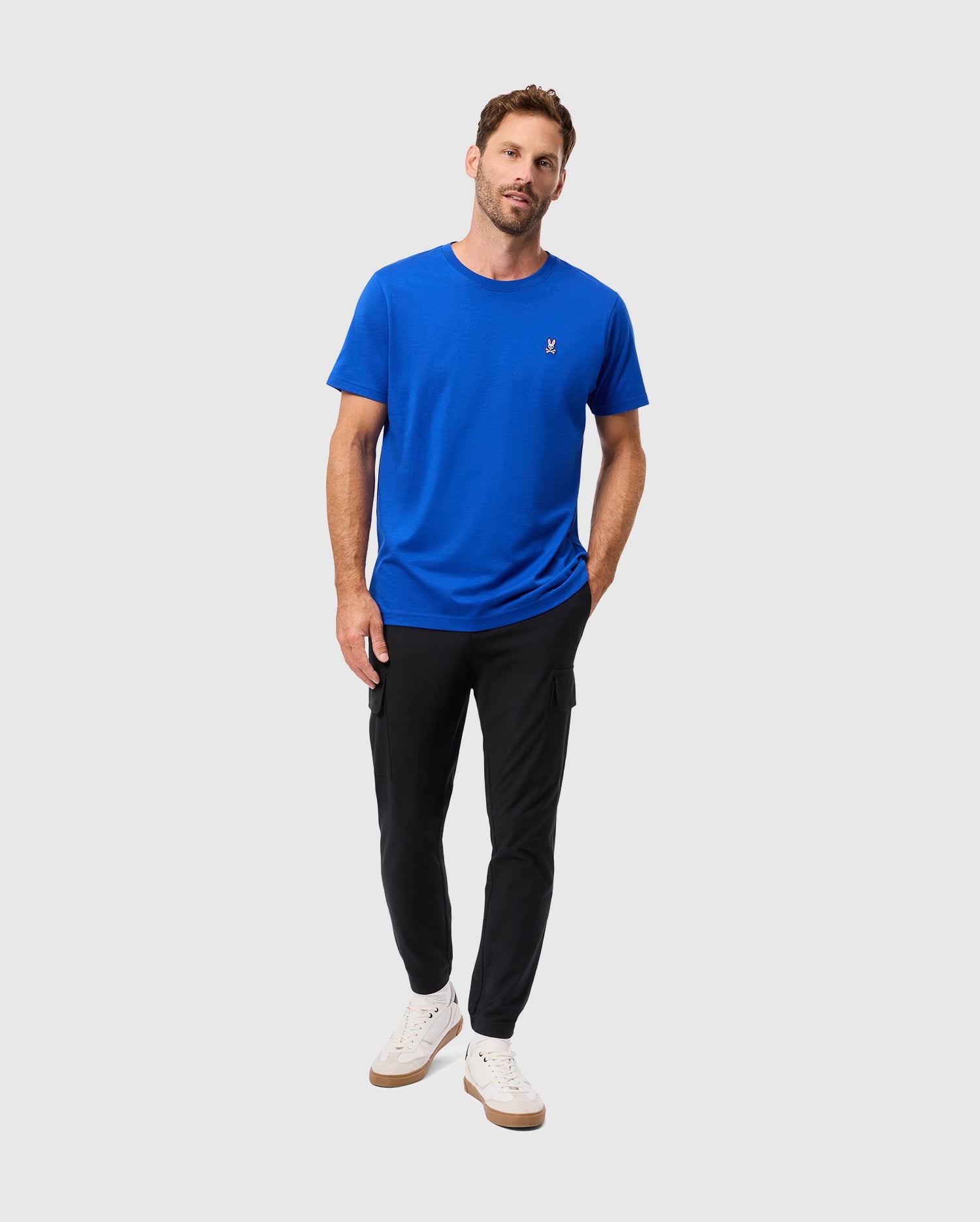 A man wearing the Psycho Bunny MENS CLASSIC CREW NECK TEE (B6U014CRPC) in blue, adorned with an embroidered Bunny logo, and black pants stands against a light gray background. He has on white sneakers and confidently looks at the camera with his hands in his pockets.