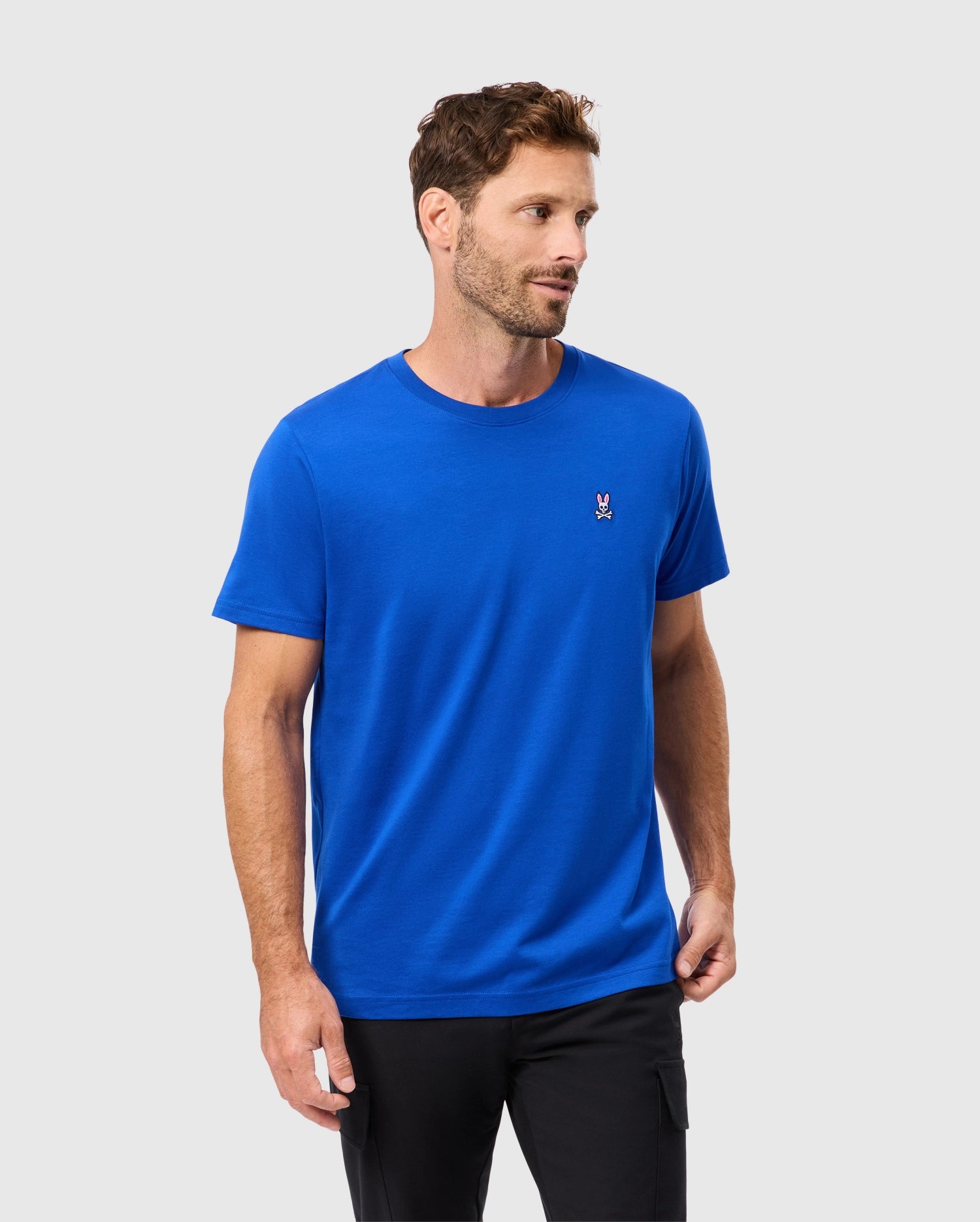 A man models the Psycho Bunny MENS CLASSIC CREW NECK TEE - B6U014CRPC in a vivid blue hue, crafted from soft Pima cotton. It features a small embroidered Bunny logo on the left side of the chest. He stands against a light gray background, looking to his left while his right hand rests in his pocket.