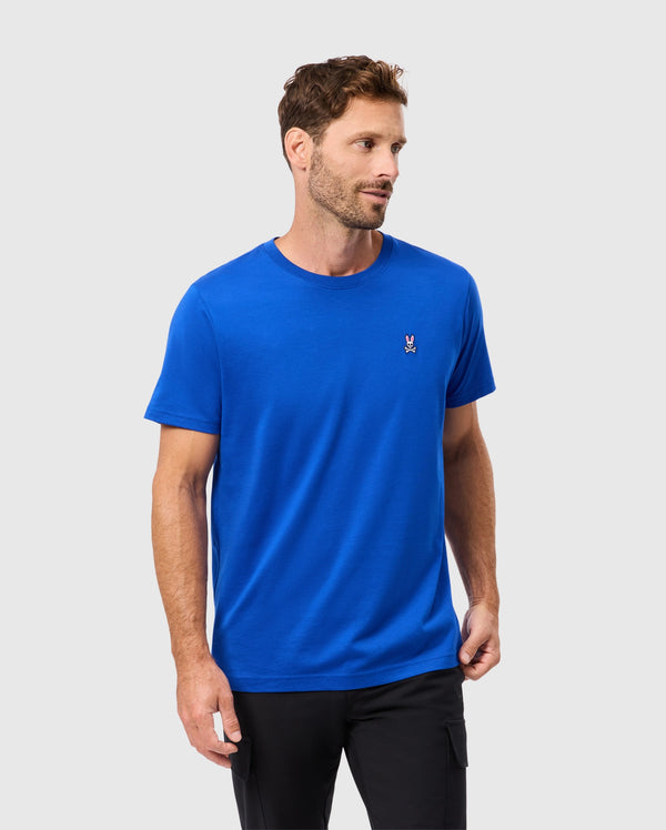 A man models the Psycho Bunny MENS CLASSIC CREW NECK TEE - B6U014CRPC in a vivid blue hue, crafted from soft Pima cotton. It features a small embroidered Bunny logo on the left side of the chest. He stands against a light gray background, looking to his left while his right hand rests in his pocket.
