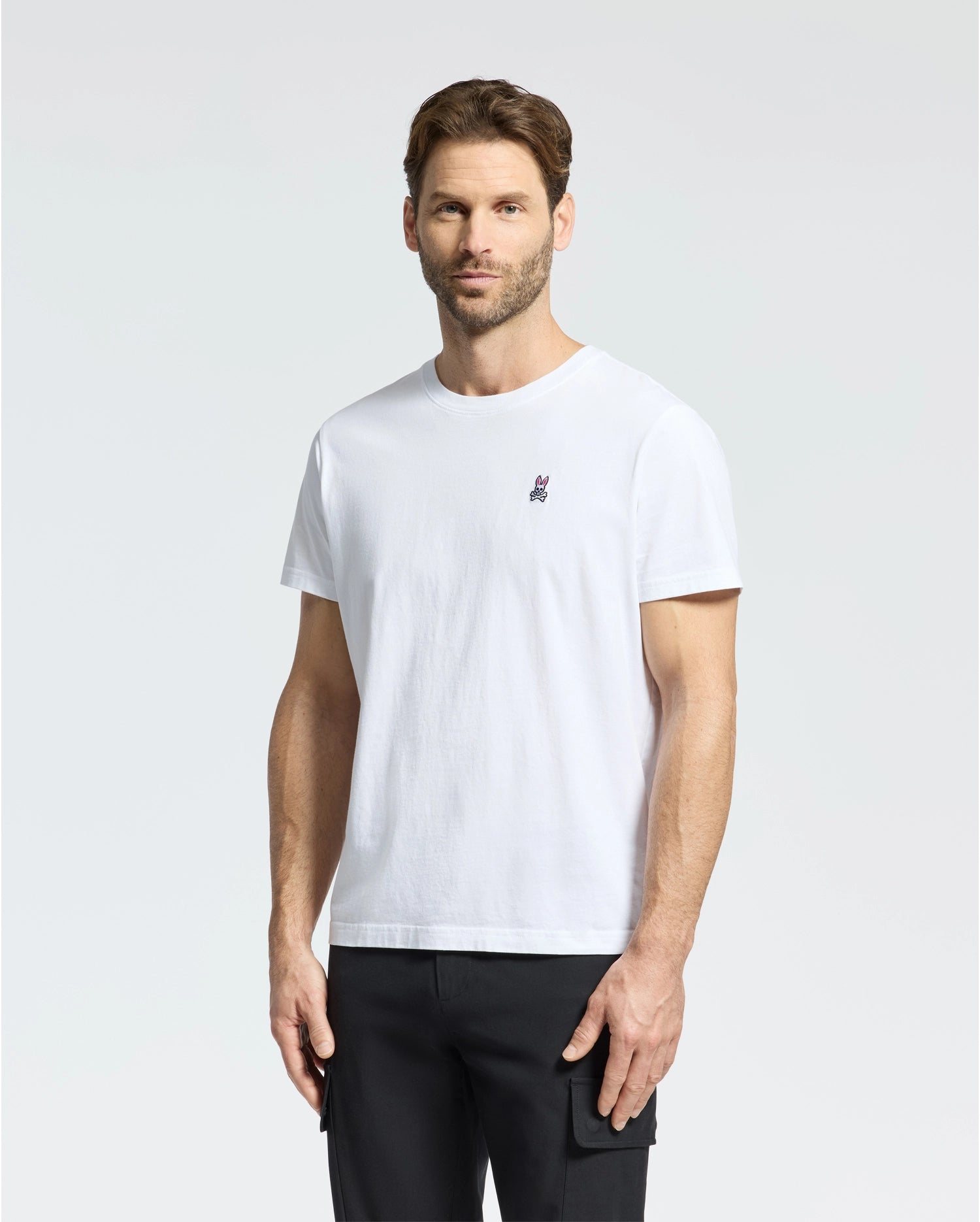 The man wears a Psycho Bunny MENS CLASSIC CREW NECK TEE - B6U014CRPC, made from 100% Pima cotton, featuring a small chest logo. He pairs it with black pants and stands facing forward on a plain light background with a neutral expression.