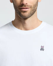 A man in a Psycho Bunny MENS CLASSIC CREW NECK TEE (B6U014CRPC), crafted from 100% Pima cotton, sports an embroidered pink rabbit and crossbones on the chest. The shirt's design stands out against the simple background, with his face partially visible.