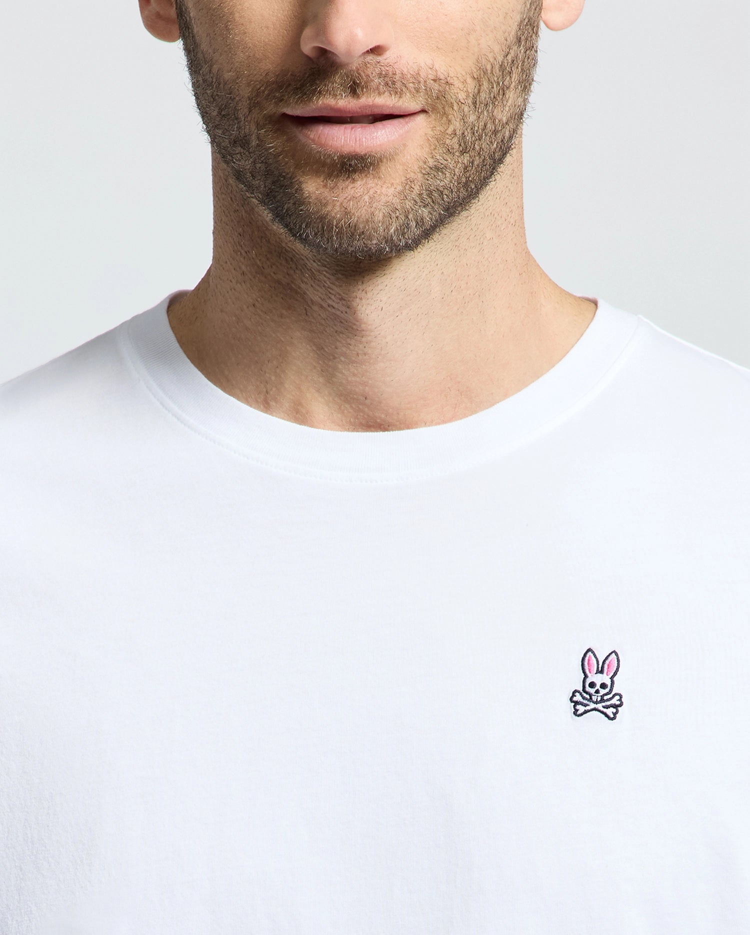 A man in a Psycho Bunny MENS CLASSIC CREW NECK TEE (B6U014CRPC), crafted from 100% Pima cotton, sports an embroidered pink rabbit and crossbones on the chest. The shirt's design stands out against the simple background, with his face partially visible.