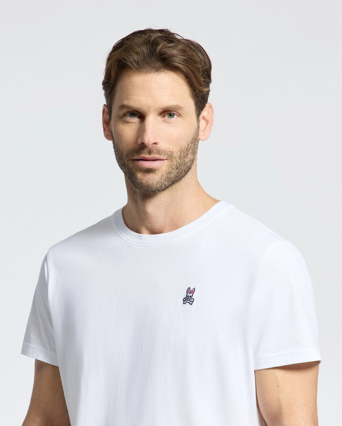 A man with short brown hair and a beard wears the Psycho Bunny MENS CLASSIC CREW NECK TEE - B6U014CRPC in 100% Pima cotton. The plain white tee features a small embroidered logo on the left. Set against a light gray backdrop, perfect for showcasing its elegant design available in Big and Tall sizes.