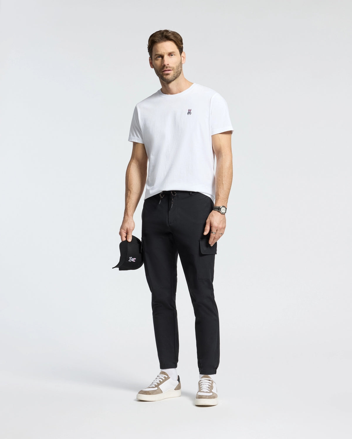 A man in a Psycho Bunny MENS CLASSIC CREW NECK TEE - B6U014CRPC made from 100% Pima cotton stands confidently in black pants, holding a black cap. He completes the look with white sneakers and a watch against a plain white background, highlighting the stylish simplicity of his Big and Tall fashion choice.