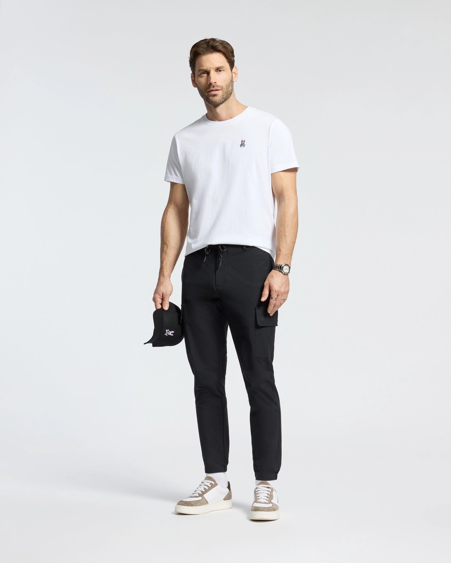 A man in a Psycho Bunny MENS CLASSIC CREW NECK TEE - B6U014CRPC made from 100% Pima cotton stands confidently in black pants, holding a black cap. He completes the look with white sneakers and a watch against a plain white background, highlighting the stylish simplicity of his Big and Tall fashion choice.