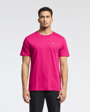A man stands against a light background in a bright pink Psycho Bunny MENS CLASSIC CREW NECK TEE - B6U014CRPC with an embroidered Bunny logo on the chest. His short hair and subtle beard complement his look, paired with sleek black pants.
