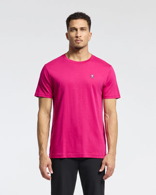 A man stands against a light background in a bright pink Psycho Bunny MENS CLASSIC CREW NECK TEE - B6U014CRPC with an embroidered Bunny logo on the chest. His short hair and subtle beard complement his look, paired with sleek black pants.