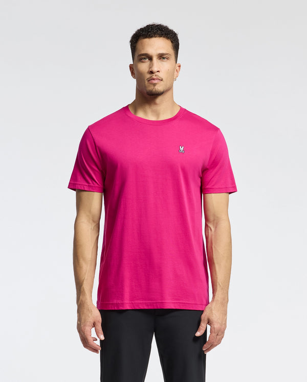 A man stands against a light background in a bright pink Psycho Bunny MENS CLASSIC CREW NECK TEE - B6U014CRPC with an embroidered Bunny logo on the chest. His short hair and subtle beard complement his look, paired with sleek black pants.