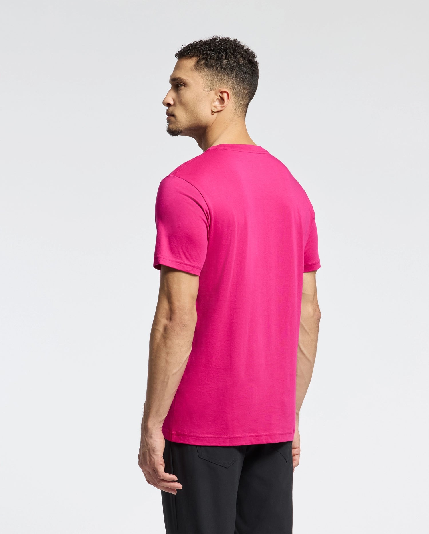 A man in a vibrant pink Psycho Bunny MENS CLASSIC CREW NECK TEE - B6U014CRPC and black pants stands sideways against a light gray backdrop, highlighting the embroidered bunny logo's subtle elegance.