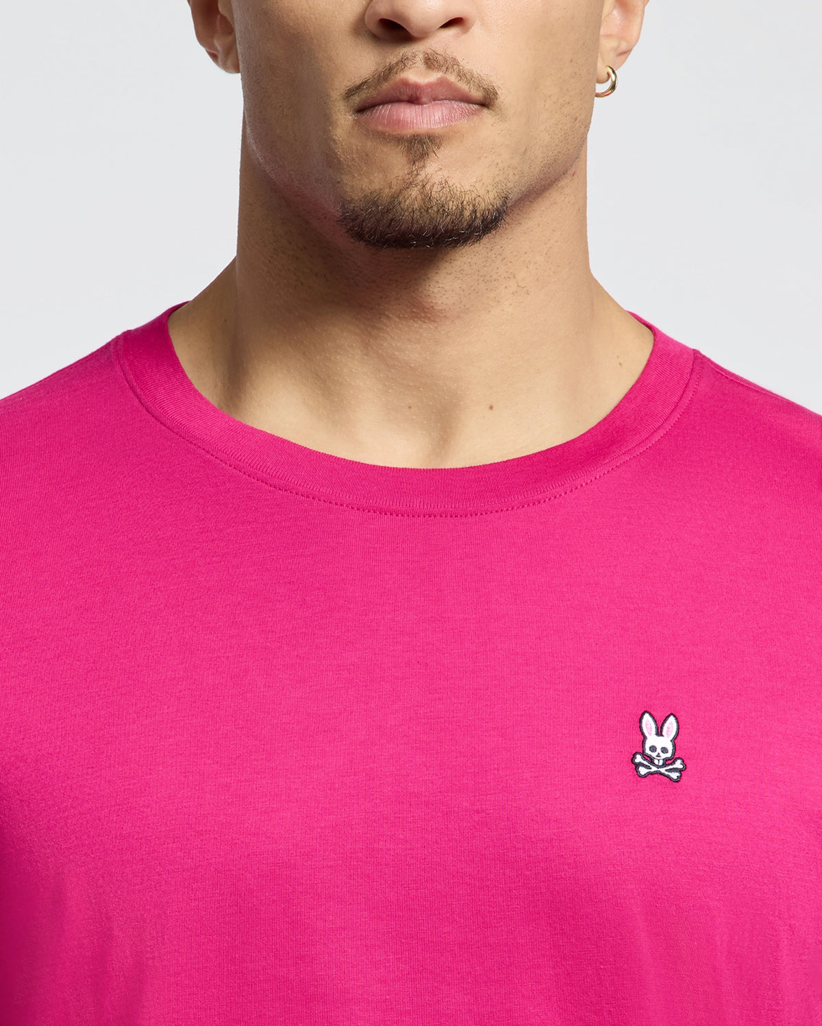 Someone is wearing a Psycho Bunny Mens Classic Crew Neck Tee in bright pink, featuring an embroidered bunny and crossbones logo on the chest. Made from luxurious Pima cotton, it stands out against a plain, light-colored background.