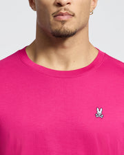 Someone is wearing a Psycho Bunny Mens Classic Crew Neck Tee in bright pink, featuring an embroidered bunny and crossbones logo on the chest. Made from luxurious Pima cotton, it stands out against a plain, light-colored background.