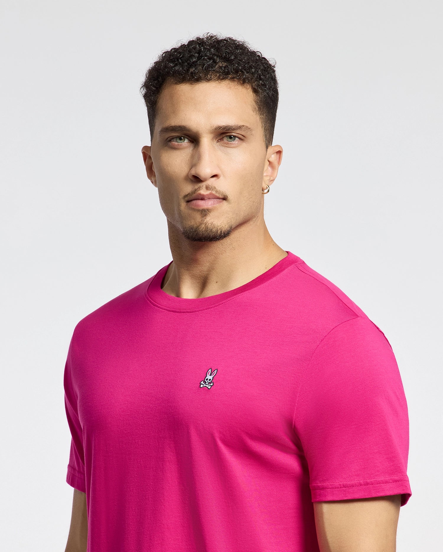 A person with short curly hair, in a Psycho Bunny MENS CLASSIC CREW NECK TEE - B6U014CRPC, vibrant pink and with an embroidered bunny logo, stands against a plain white background. They wear gold hoop earrings and have a neutral expression.
