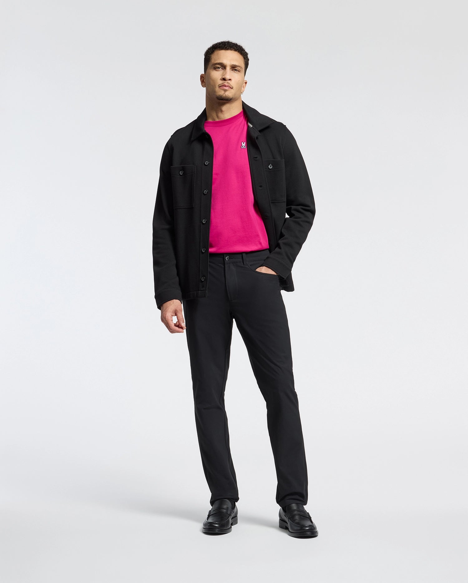 A man with short hair and a beard, standing against a plain background, wears a bright pink Psycho Bunny Classic Crew Neck Tee (B6U014CRPC) under a black jacket with matching pants and shoes. One hand in his pocket complements the ensemble, highlighted by subtle embroidered Bunny logo details.