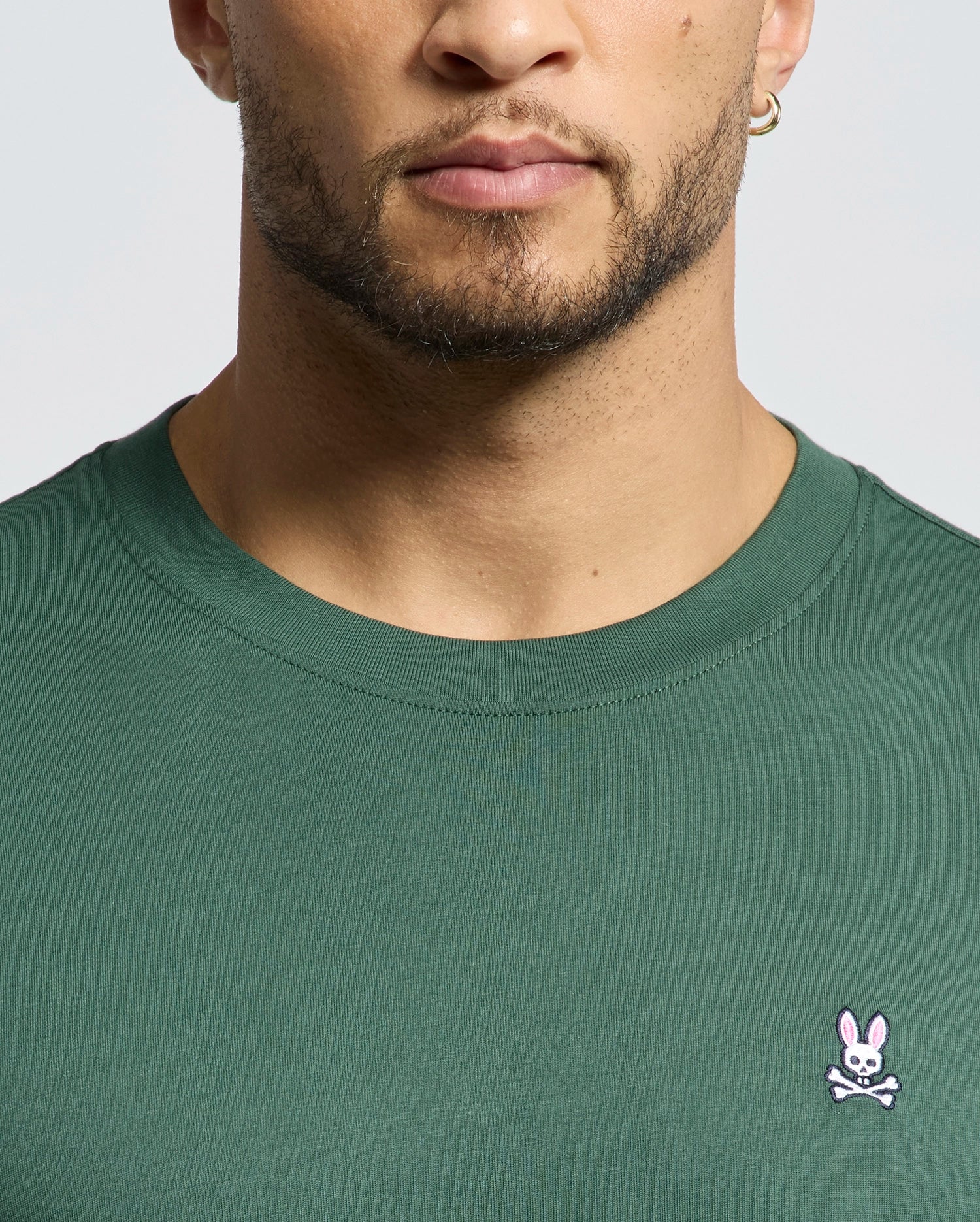 A man wears the Psycho Bunny MENS CLASSIC CREW NECK TEE (B6U014F200) in green, featuring a small pink bunny and white crossbones logo on the left chest. Only his lower face, neck, and upper chest are shown.