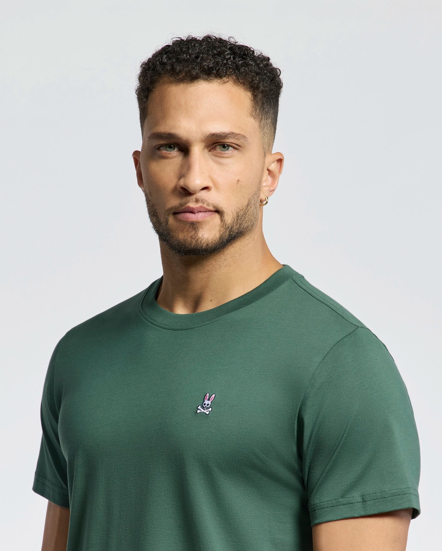 A man with short curly hair and a trimmed beard wears the Psycho Bunny MENS CLASSIC CREW NECK TEE (B6U014F200) in green, featuring a small bunny logo on the chest, standing against a plain light background.