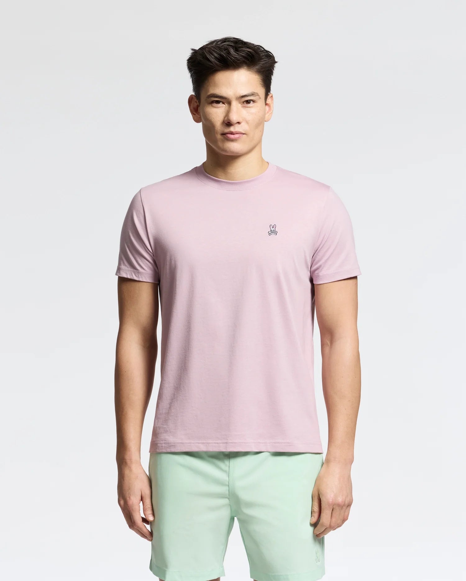 Wearing a Psycho Bunny MENS CLASSIC CREW NECK TEE in light pink, made from 100% pima cotton with a chest logo, paired with mint green shorts, the person stands against a white background.