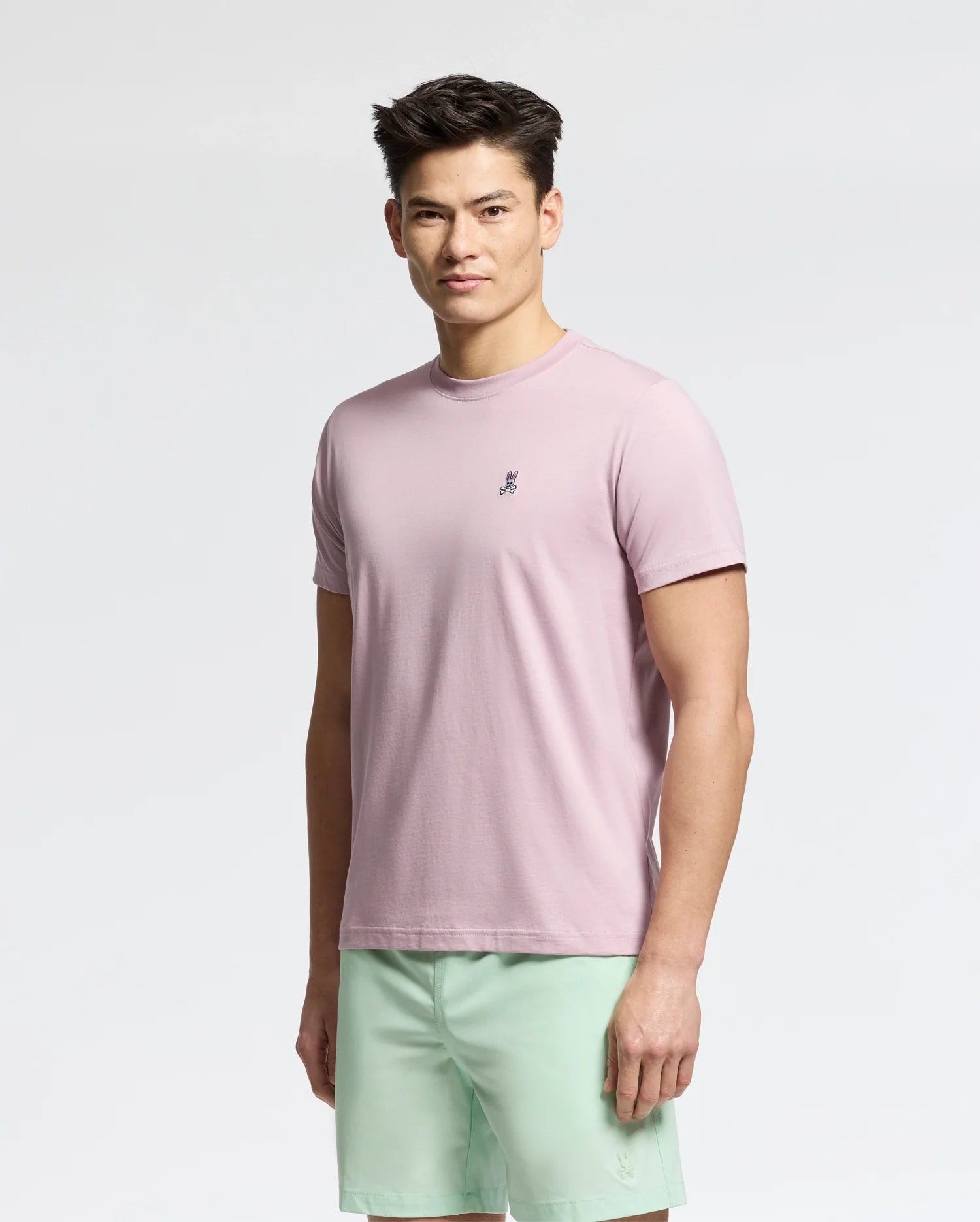 In front of a plain white background, a person with short dark hair and a neutral expression wears Psycho Bunny's MENS CLASSIC CREW NECK TEE - B6U014F200 in light pink, made from 100% pima cotton, paired with mint green shorts.