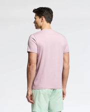 A man faces away from the camera, highlighting a Psycho Bunny MENS CLASSIC CREW NECK TEE - B6U014F200 in light pink with light green shorts. Crafted from 100% pima cotton, this versatile attire stands out against a plain white backdrop.
