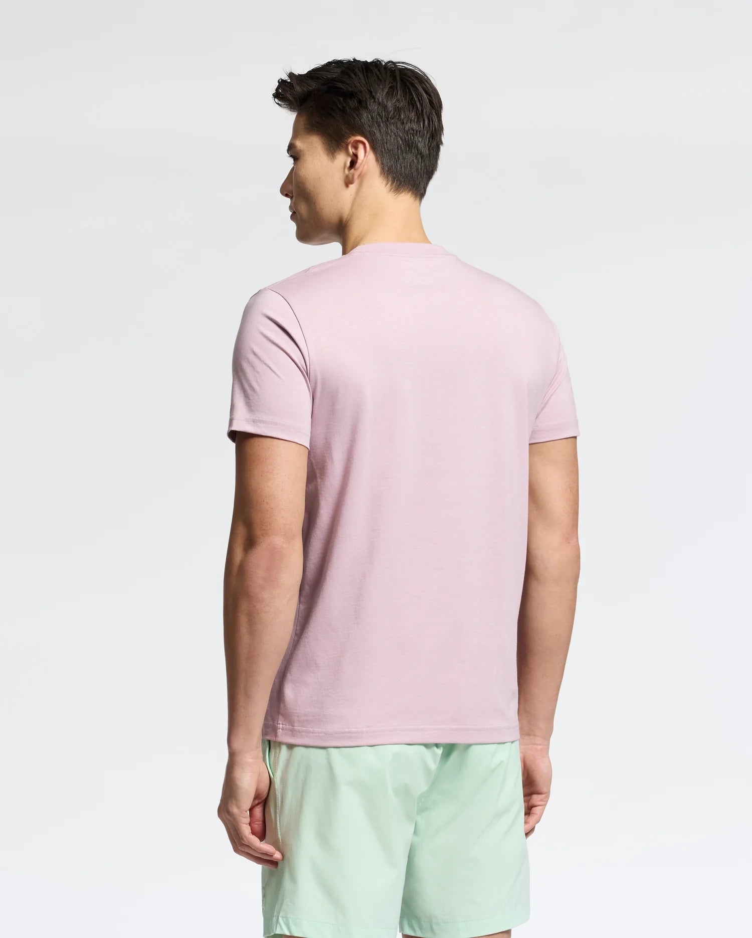 A man faces away from the camera, highlighting a Psycho Bunny MENS CLASSIC CREW NECK TEE - B6U014F200 in light pink with light green shorts. Crafted from 100% pima cotton, this versatile attire stands out against a plain white backdrop.