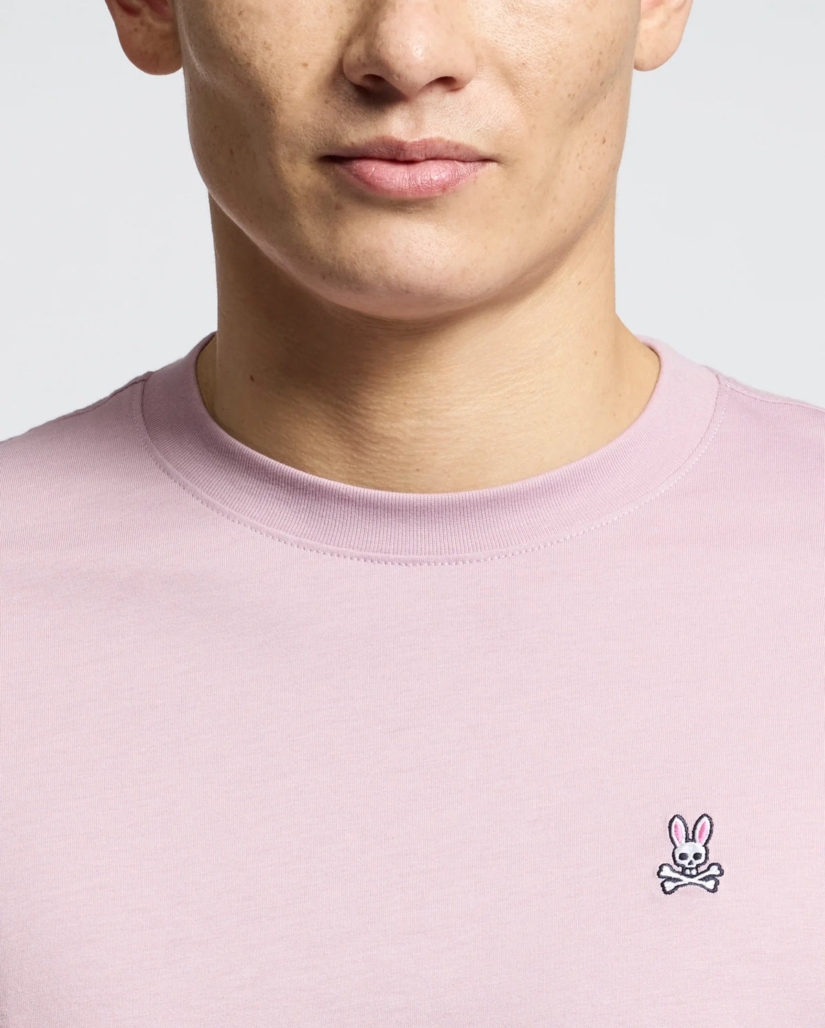 A person wearing the Men's Classic Crew Neck Tee in light pink from Psycho Bunny demonstrates its versatility, featuring a small embroidered bunny with crossed bones logo on the chest. This 100% pima cotton tee highlights their upper torso against a neutral background.