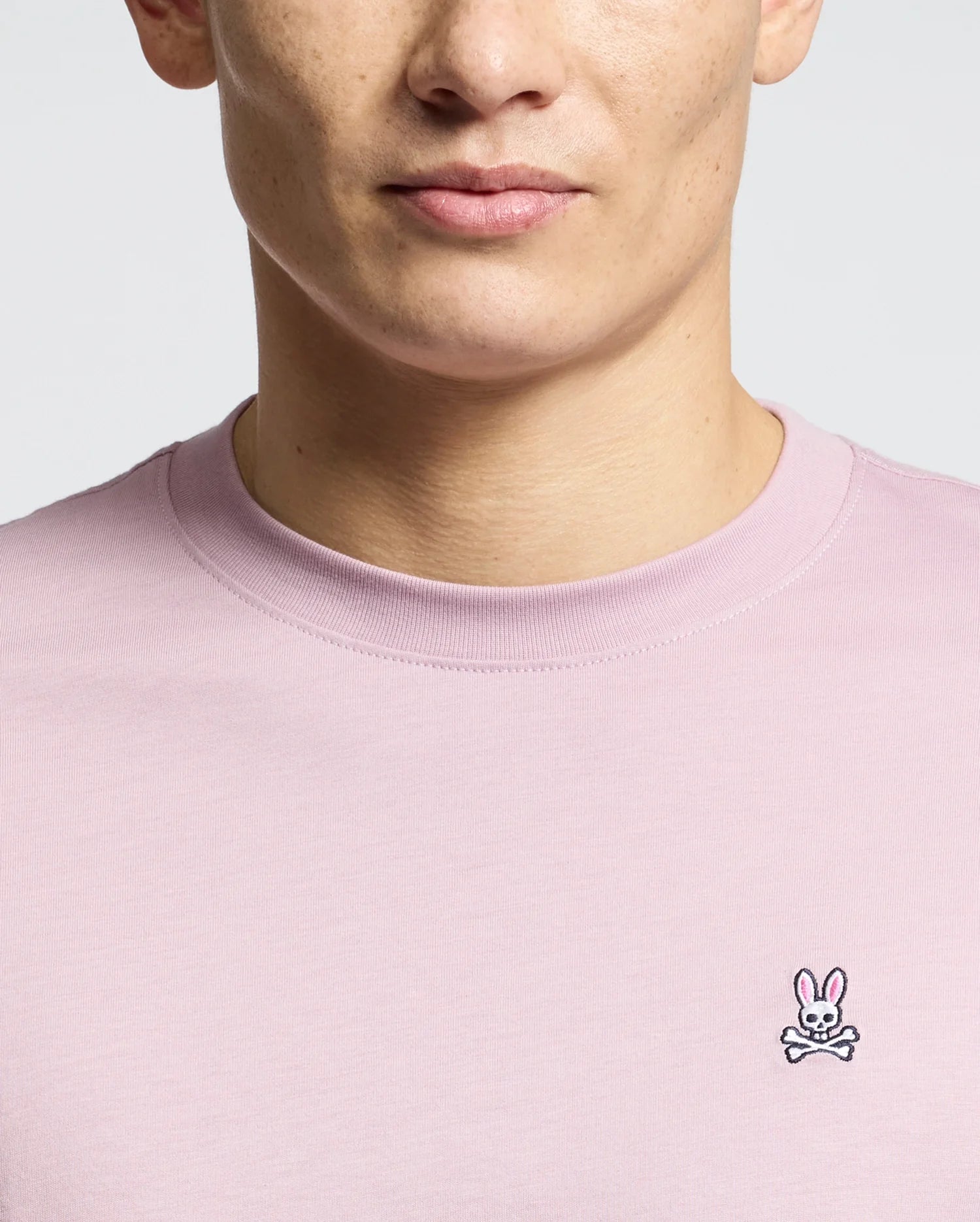 A person wearing the Men's Classic Crew Neck Tee in light pink from Psycho Bunny demonstrates its versatility, featuring a small embroidered bunny with crossed bones logo on the chest. This 100% pima cotton tee highlights their upper torso against a neutral background.