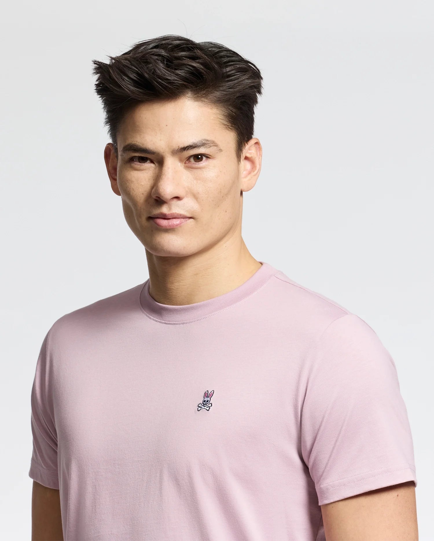 A person in a versatile light pink Psycho Bunny MENS CLASSIC CREW NECK TEE - B6U014F200, made from 100% pima cotton and featuring a small embroidered rabbit on the chest, stands against a plain white background with short styled dark hair and a neutral expression facing the camera.