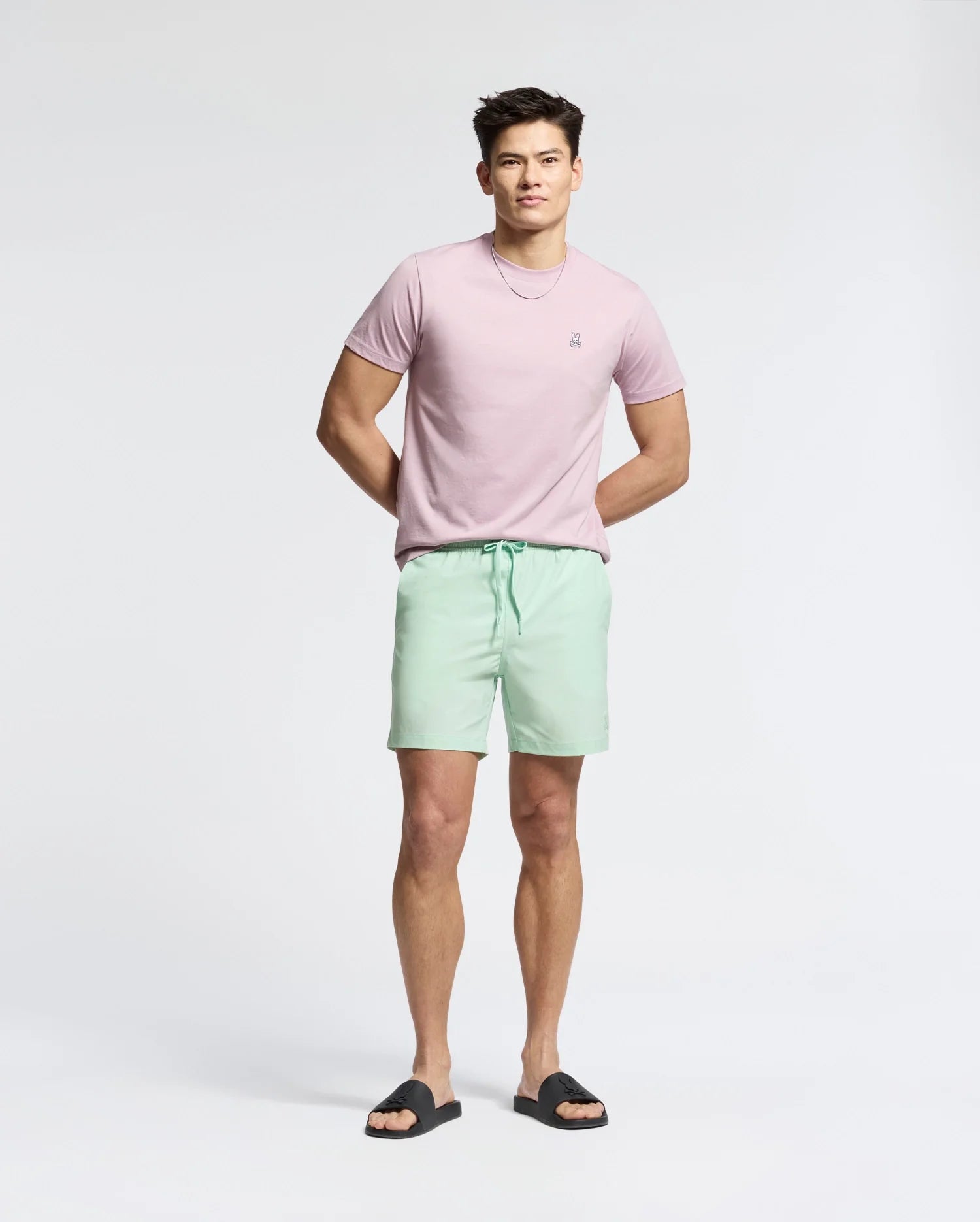 A man is wearing a Psycho Bunny light pink MENS CLASSIC CREW NECK TEE - B6U014F200 made of 100% pima cotton, paired with mint green shorts and black sandals. His hands are loosely clasped behind his back, creating a versatile look on a plain white background.
