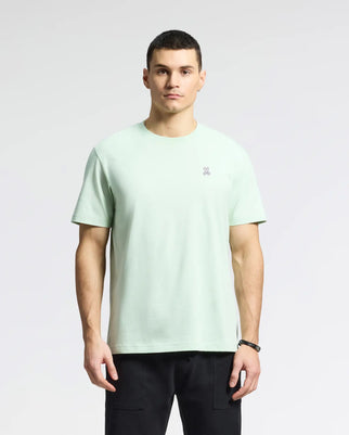 A person wears the Men's Classic Crew Neck Tee by Psycho Bunny in light green, made of soft Pima cotton with a subtle Bunny logo on the chest, against a plain gray background and paired with sleek black pants.