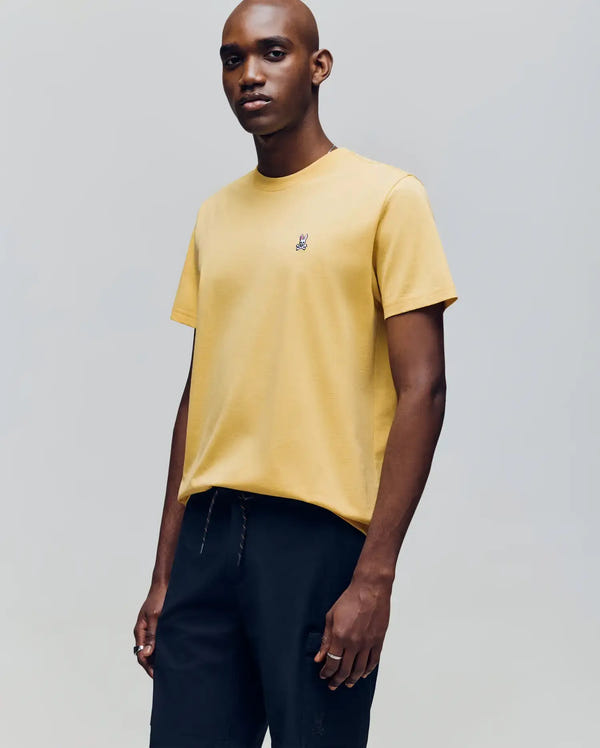 A man wears a Psycho Bunny MENS CLASSIC CREW NECK TEE (B6U014F200) in yellow, featuring a small chest logo, paired with black pants, standing against a plain light grey background.