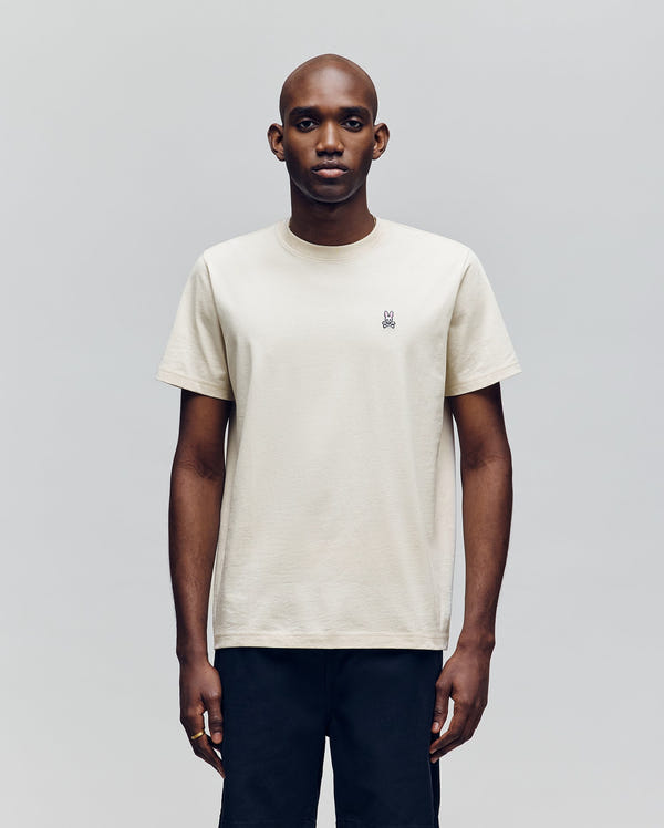 A man faces forward against a light gray background, wearing Psycho Bunny's MENS CLASSIC CREW NECK TEE - B6U014F200, a regular-fit, short-sleeved off-white pima cotton shirt with an embroidered chest design and dark pants. His expression is neutral.