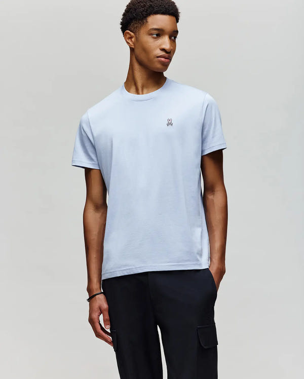 A man stands against a light gray background wearing a Psycho Bunny MENS CLASSIC CREW NECK TEE (B6U014F200) with a small bunny logo on the chest and black cargo pants.