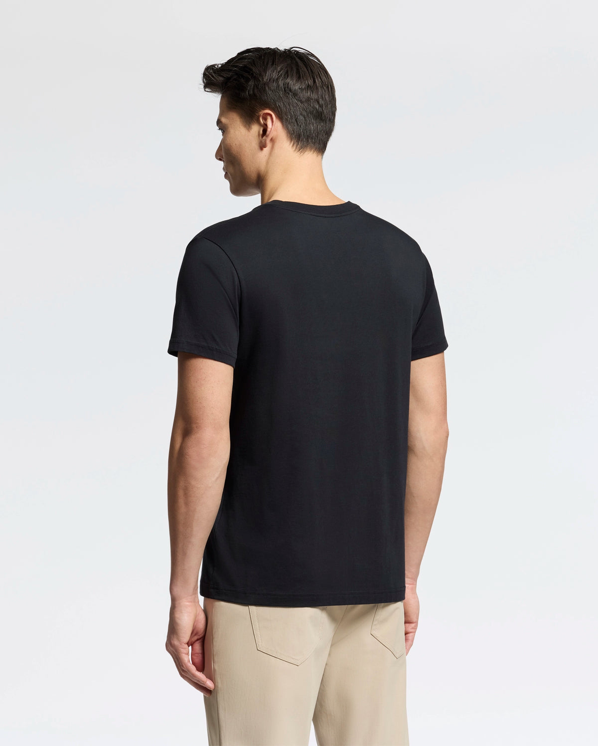A man faces away from the camera, dressed in a plain black Psycho Bunny Men's Classic V Neck Tee (B6U100ARPC) and beige pants against a simple white background.