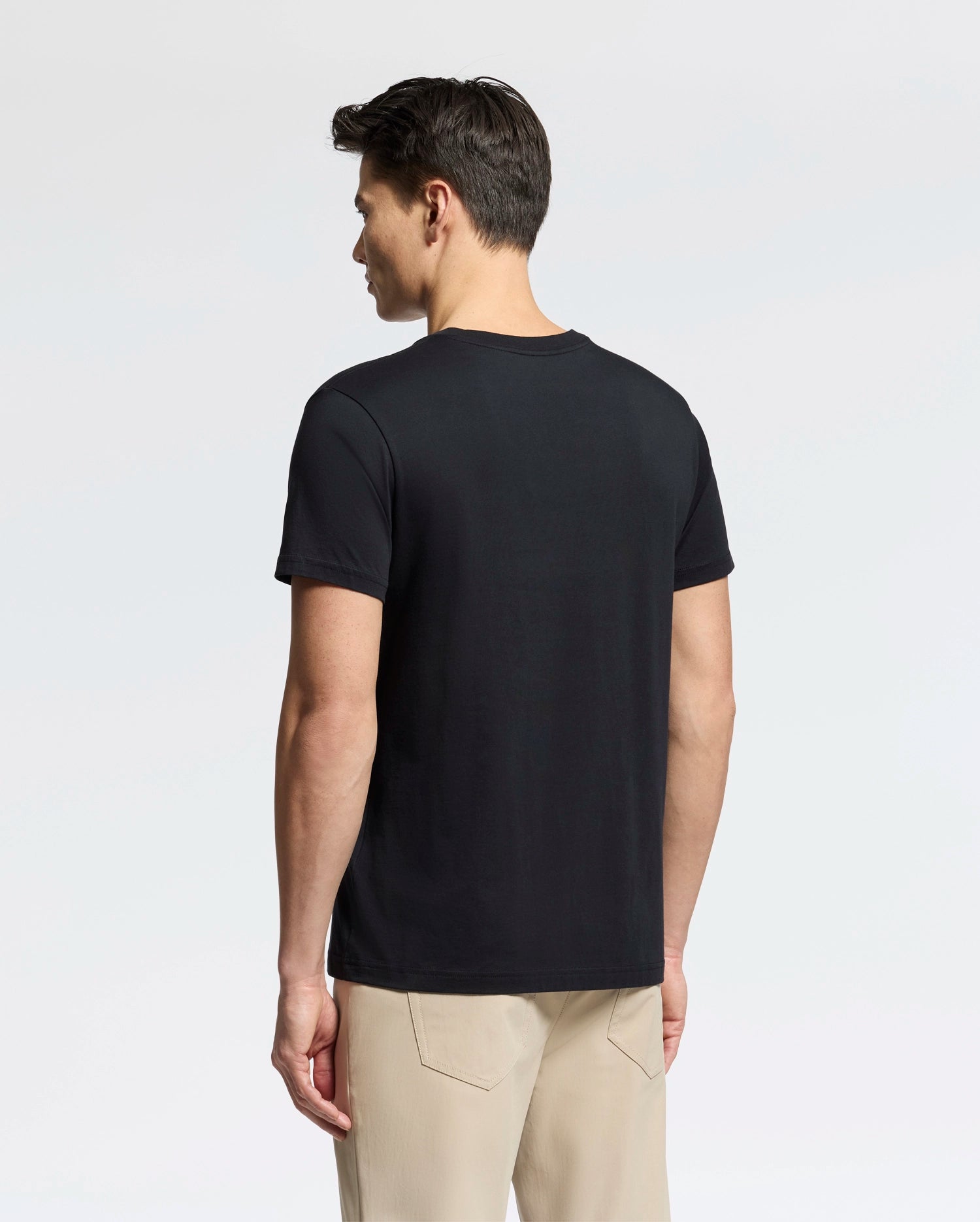 A man faces away from the camera, dressed in a plain black Psycho Bunny Men's Classic V Neck Tee (B6U100ARPC) and beige pants against a simple white background.
