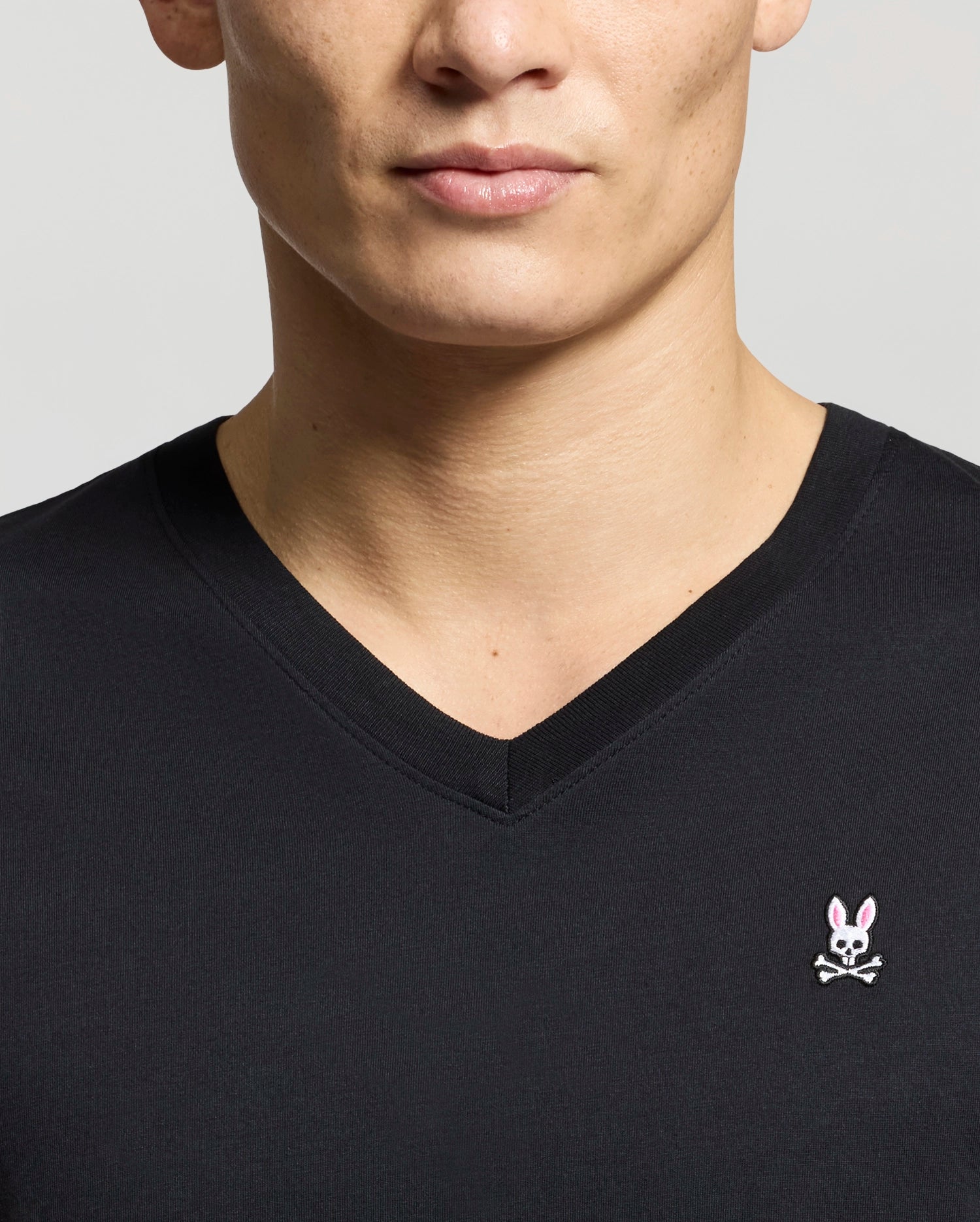 A person is wearing a Psycho Bunny Men's Classic V-Neck Tee (B6U100ARPC) in black, showcasing the pink bunny head and crossbones logo on the left side. The image is cropped to display only the upper torso and part of the face.