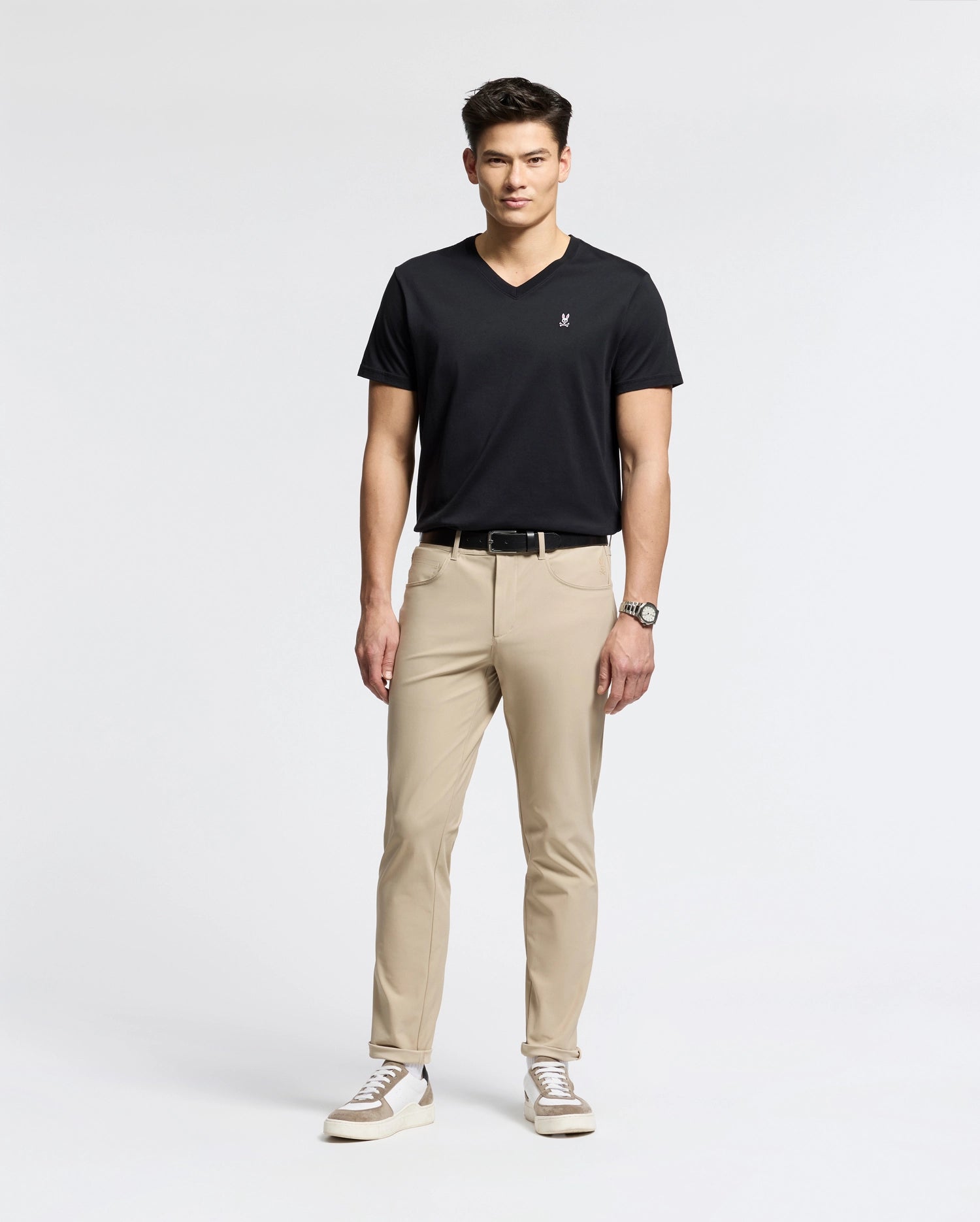 A man exudes confidence in a sleek black Psycho Bunny MENS CLASSIC V NECK TEE - B6U100ARPC, crafted from soft Pima cotton. Paired with beige pants and white sneakers, he embodies effortless style, his wrist adorned with a watch as he gazes forward against a plain background.