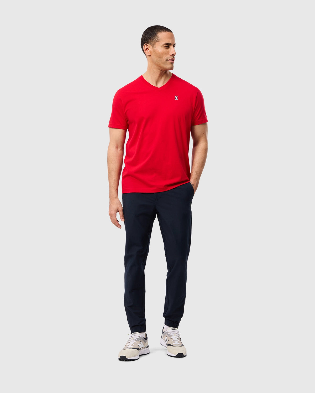 A man is standing against a plain background, wearing the Psycho Bunny Men's Classic V Neck Tee (B6U100CRPC) in red, navy blue pants, and white sneakers. He has short hair and is looking to his left with his right hand in his pocket and his left hand by his side. The tee, which features the Psycho Bunny logo, is made of 100% Pima cotton for ultimate comfort.