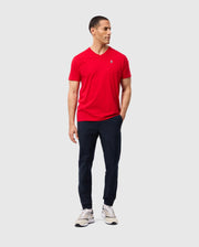 A man is standing against a plain background, wearing the Psycho Bunny Men's Classic V Neck Tee (B6U100CRPC) in red, navy blue pants, and white sneakers. He has short hair and is looking to his left with his right hand in his pocket and his left hand by his side. The tee, which features the Psycho Bunny logo, is made of 100% Pima cotton for ultimate comfort.