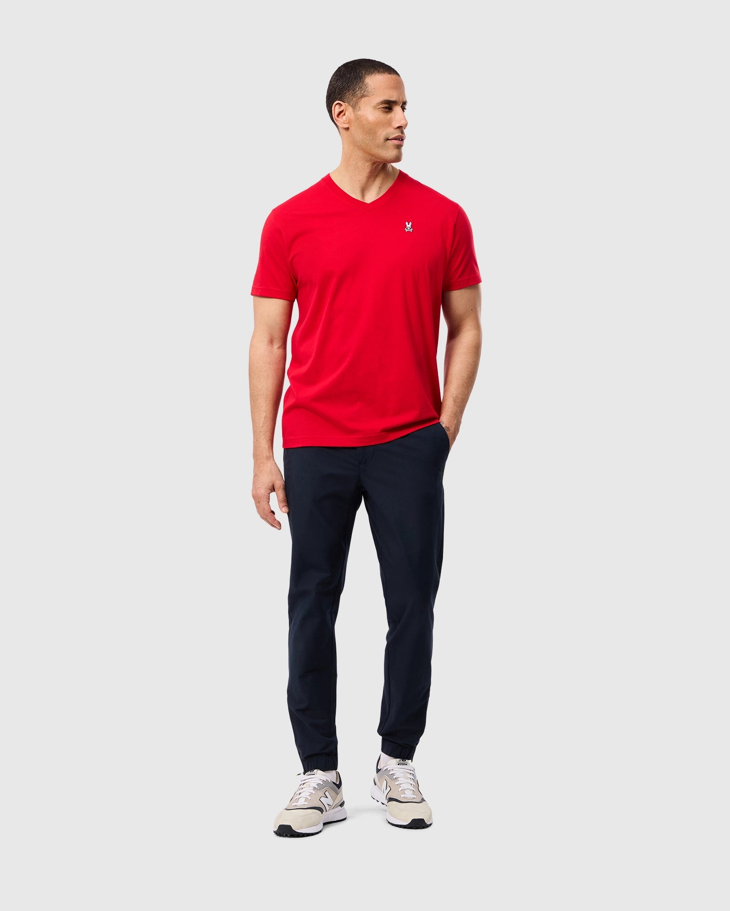 A man is standing against a plain background, wearing the Psycho Bunny Men's Classic V Neck Tee (B6U100CRPC) in red, navy blue pants, and white sneakers. He has short hair and is looking to his left with his right hand in his pocket and his left hand by his side. The tee, which features the Psycho Bunny logo, is made of 100% Pima cotton for ultimate comfort.